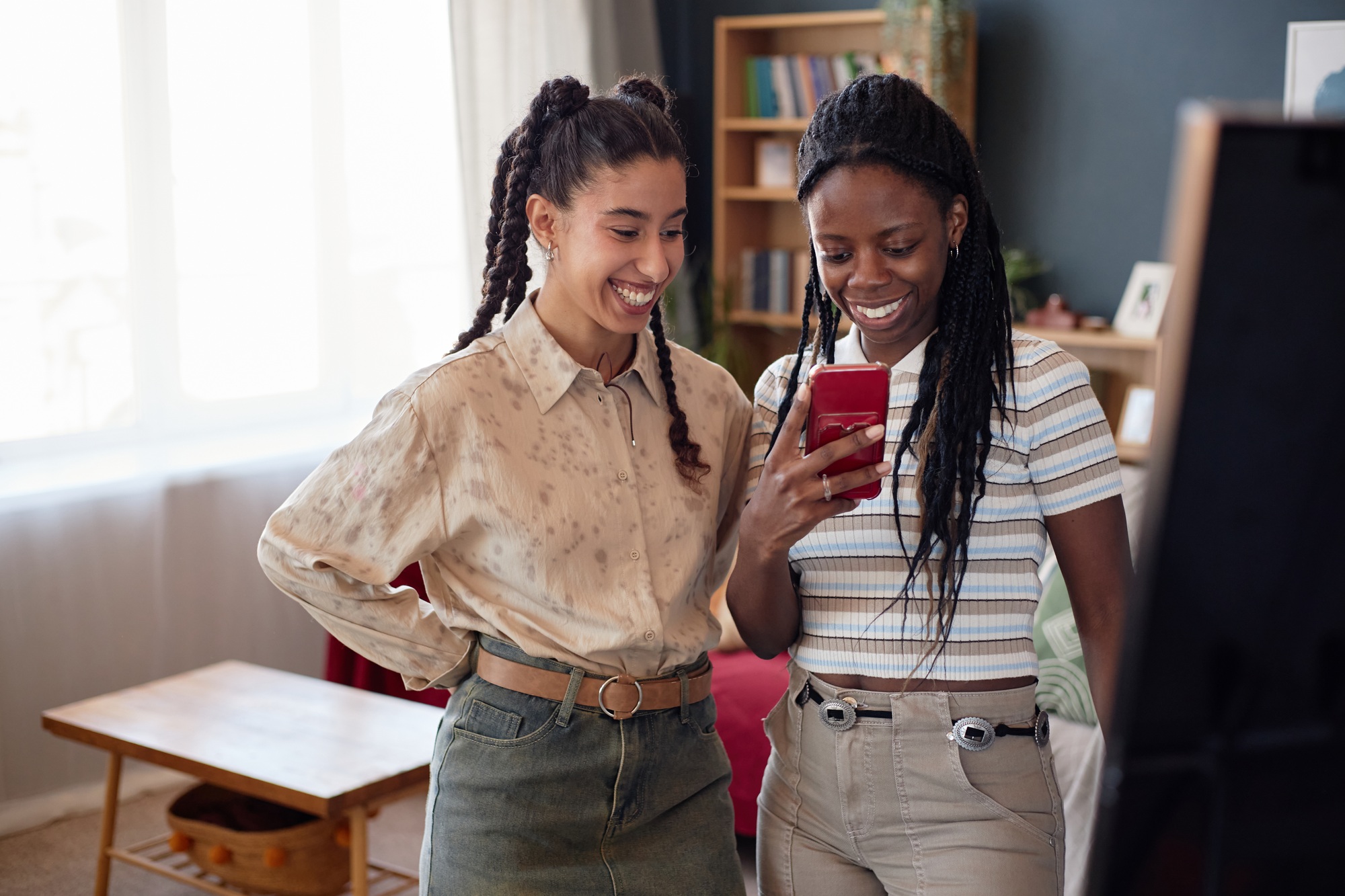 Two Young Adult Multiethnic Women Smiling While Looking at Smartphone Indoors
